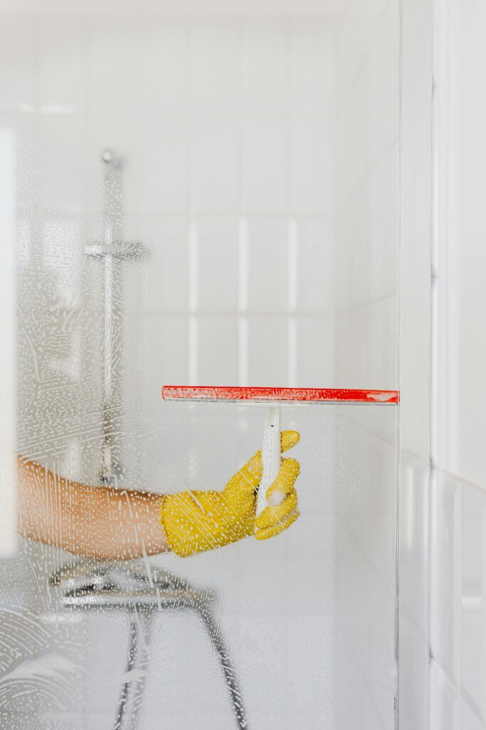 Crop anonymous person in yellow rubber glove cleaning glass shower unit with glass wiper and detergent against white tile wall